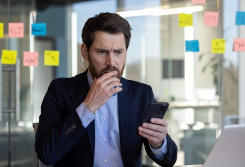 Professional businessman in a modern office setting focused on his smartphone, displaying a concerned expression with sticky notes in the background.