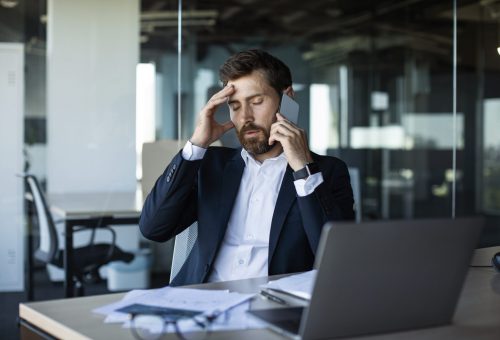 Concerned male entrepreneur talking on cellphone and touching head in stress, worried middle aged businessman suffering problems with work, having unpleasant mobile call