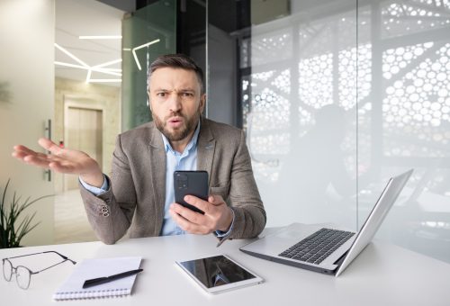 Confused businessman with phone at desk expressing frustration, surrounded by tech gadgets in a modern office setting.